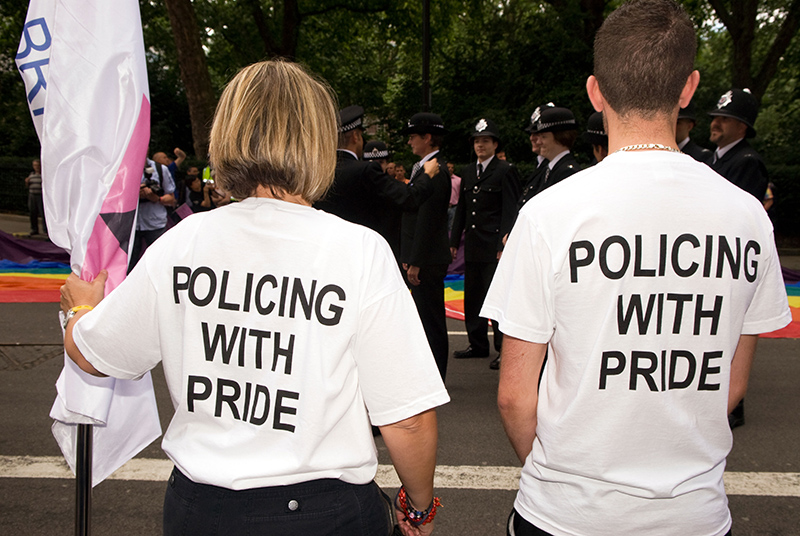 Two individuals stand together, wearing white shirts that read "Policing with Pride." They support LGBTQ+ inclusivity alongside police officers at a vibrant, community-oriented event.