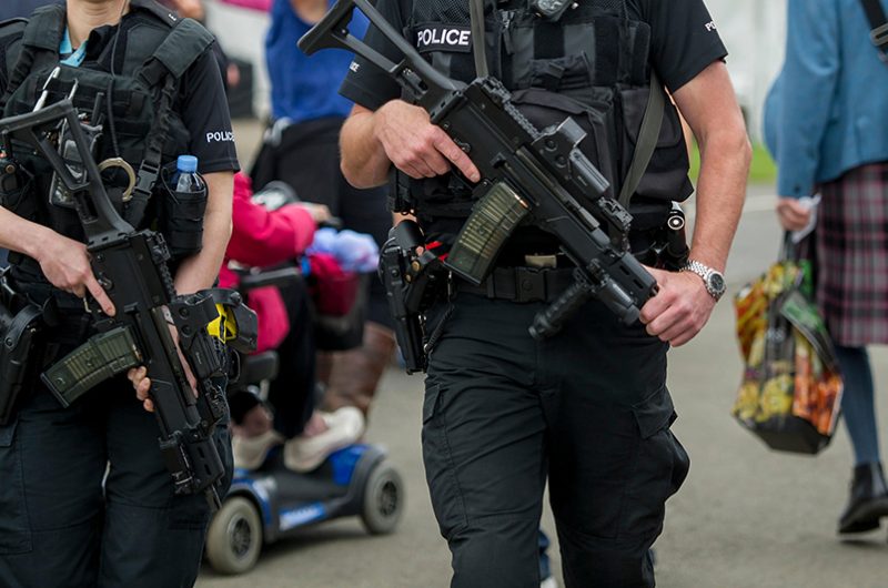 Two police officers, (faces cropped out) equipped with tactical gear and rifles, walk through a crowd, emphasizing their readiness and presence in maintaining public safety within the community.