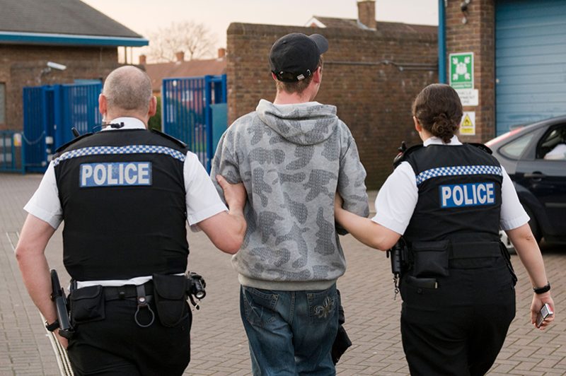 Two police officers are escorting a person in handcuffs and a hoodie towards a police station, indicating an arrest or detainment in a professional law enforcement context.