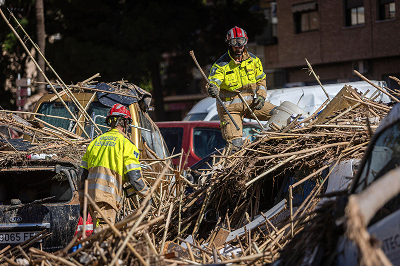 Rescue workers in bright jackets navigate a chaotic scene of debris, removing branches and rubble from damaged vehicles after a disaster, exemplifying emergency response efforts.