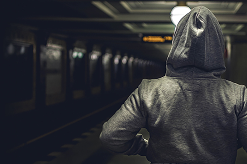 Young person pictured from behind in hoodie, standing in a dark railway station