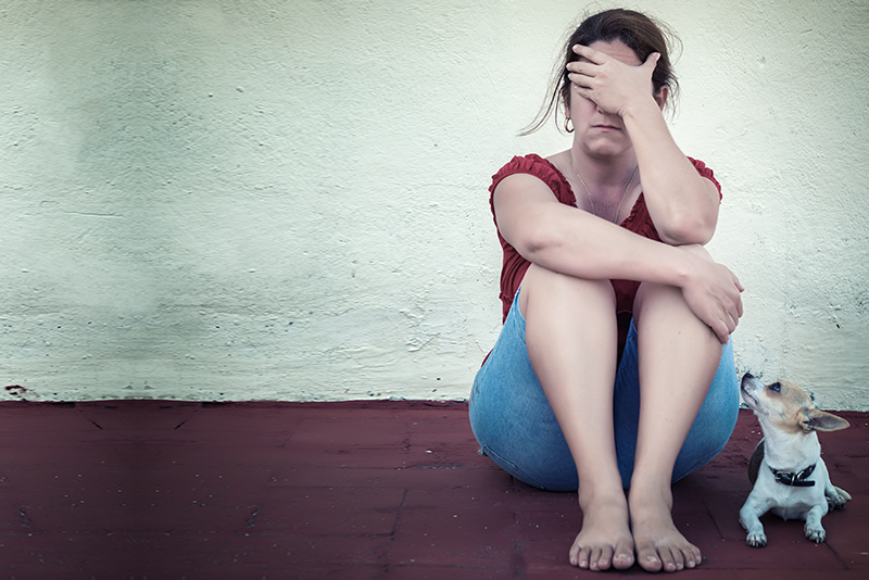 Upset women, victim of crime, sits against a wall with her hand over her face, and her dog alongside her