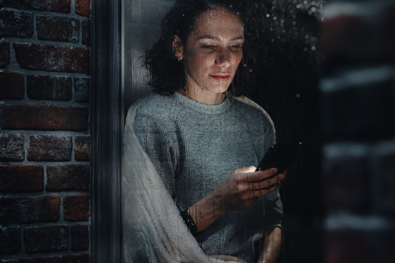 A woman, wrapped in a cozy blanket, gazes at her smartphone, illuminated by soft light, while rain gently streams down the window beside her, creating a serene atmosphere.