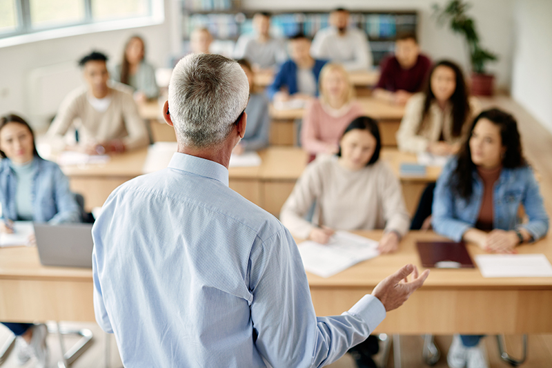 A teacher stands at the front of a classroom, engaging several attentive students who are seated at desks, creating an atmosphere of learning and discussion on policing insights.