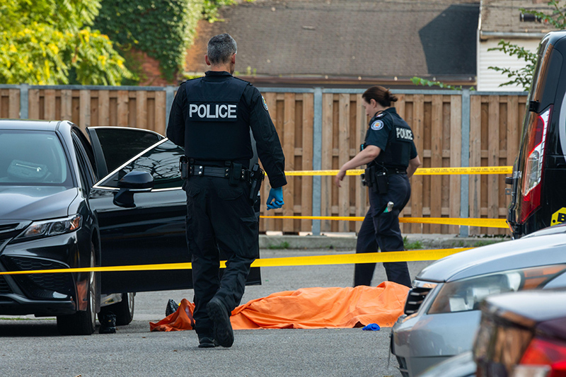 Two police officers investigate a fatal shooting in Toronto. An orange tarp covers an object on the ground, indicating a serious incident has occurred. The atmosphere feels tense and somber.