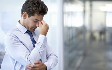 A man in a light blue shirt looks stressed, rubbing his forehead in an office setting, highlighting the pressures often faced in policing and related professions.