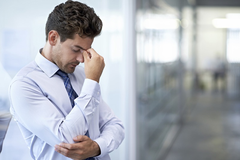 A man in a light blue shirt looks stressed, rubbing his forehead in an office setting, highlighting the pressures often faced in policing and related professions.