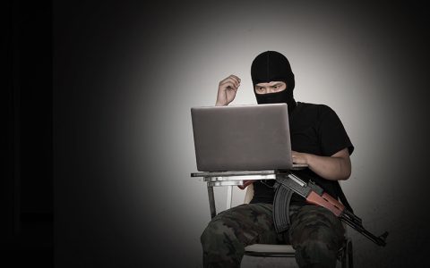 Man with a hood, combat trousers and automatic weapon sitting in a. darkened room on a laptop – online terrorist content