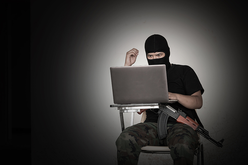 Man with a hood, combat trousers and automatic weapon sitting in a. darkened room on a laptop – online terrorist content
