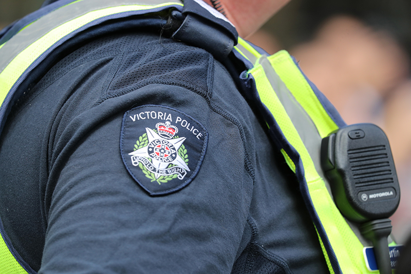 The image features a police officer's uniform, highlighting the "Victoria Police" badge on the shoulder, with reflective yellow details and a communication device attached, symbolizing authority and readiness.