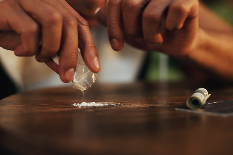 Person's hands tipping cocaine onto a table top, money rolland credit card nearby