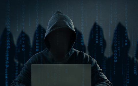 Hooded hacker sitting in a dark room at a laptop, with the silhouettes of other hackers behind him