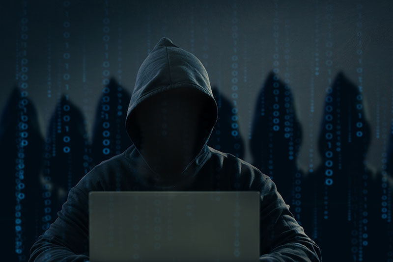Hooded hacker sitting in a dark room at a laptop, with the silhouettes of other hackers behind him