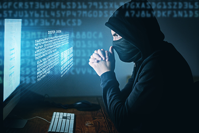 Man in hoodie and face mask sitting in front of a computer screen with lots of coding; cybercrime or hacker