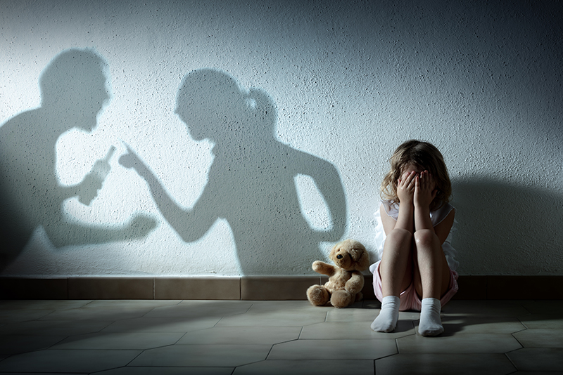 Young girl sits on the floor against a wall, covering her eyes, as the shadows of her parents fighting can be seen on the wall beside her