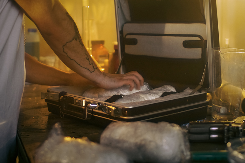 An individual is carefully packing a suitcase with tightly wrapped bundles, of drugs surrounded by laboratory equipment and containers