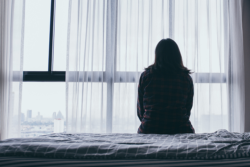 Woman sitting on her own on a bed, looking out of a window, in silhouette – victim of domestic violence or other crime