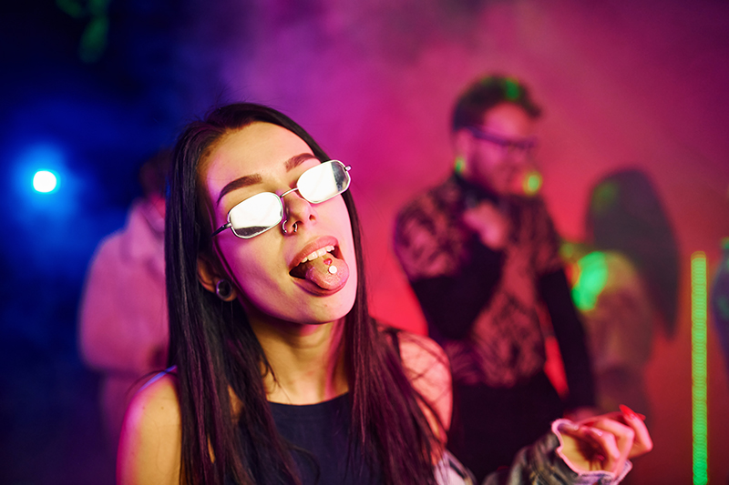 Young girl holding drug pills on togue in front of young people that having fun in night club with colorful laser lights.