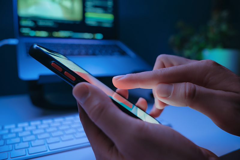 Close-up of persons hand taping on smartphone screen with social media feed at night with laptop on background.