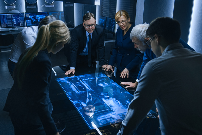 Team of Government Intelligence / FBI Agents Standing Around Digital Touch Screen Table and Tracking Suspect Vehicle Using Satellite Surveillance in the Monitoring Room.