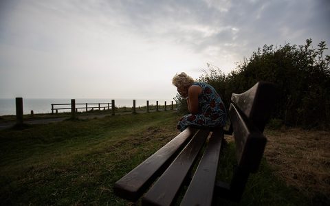 silhouetted mature woman coastal bench scene, isolated seated figure near fenced cliff edge,