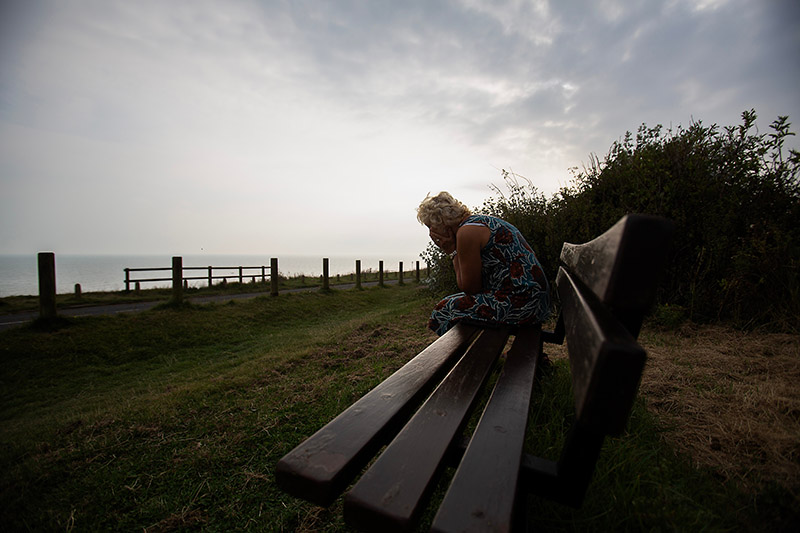 silhouetted mature woman coastal bench scene, isolated seated figure near fenced cliff edge,