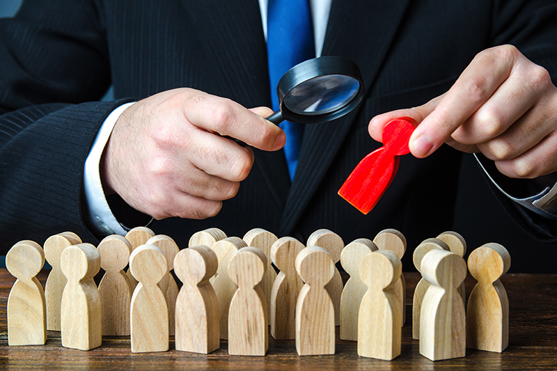 A suited figure examines wooden people with a magnifying glass, highlighting one red figure among many, symbolizing scrutiny and focus in policing or investigations.