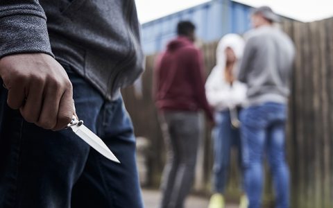 A person grips a knife, standing close to others engaged in conversation, suggesting a tense situation. The setting is an outdoor area with a wooden fence and containers nearby.