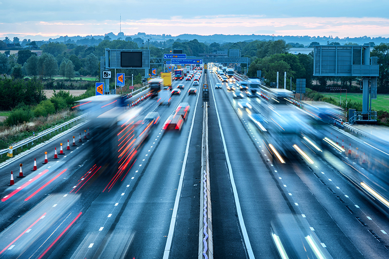A busy highway with multiple lanes, featuring blurred lights from speeding vehicles and surrounding construction cones, illustrating traffic flow and potential monitoring for safety enforcement.