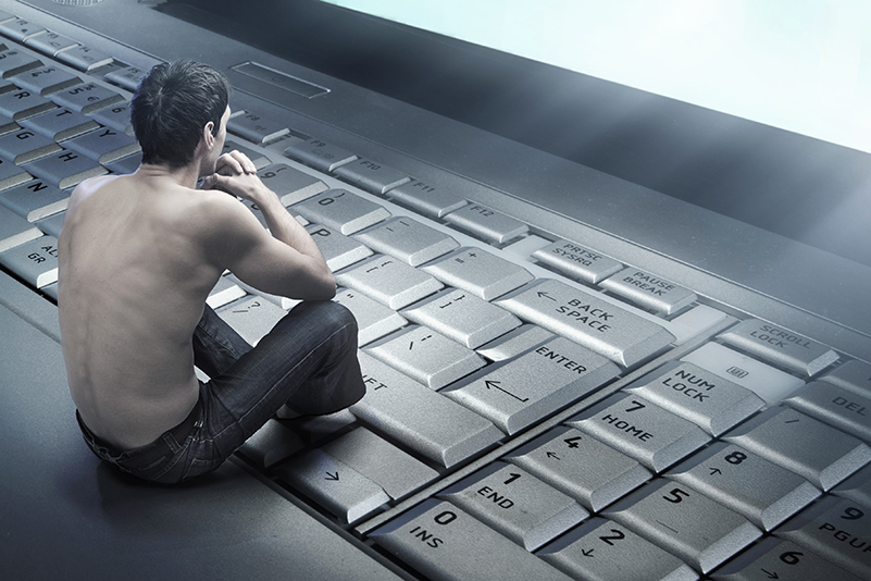 Conceptual photo of a young man sitting on a large laptop keyboard staring at the screen, addicted to the internet, dangers of online extremism misogyny radicalisation