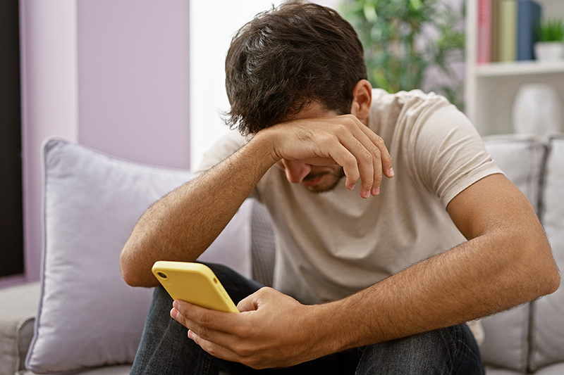 A young hispanic man with a beard looks distressed while using a smartphone in a modern living room setting.