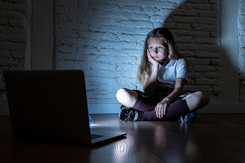 young girl sitting in front of a laptop in a dark room, online abuse CSAM