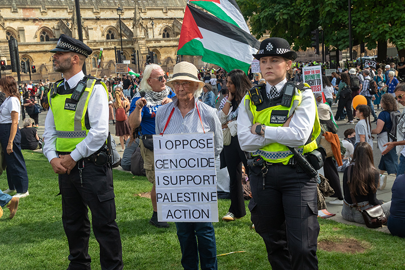 Pro-Palestine Protesters in Parliament Square hold signs reading I oppose Genocide, I support Palestine Action, with one elderly female protestors flanked either side by a male and female police officer