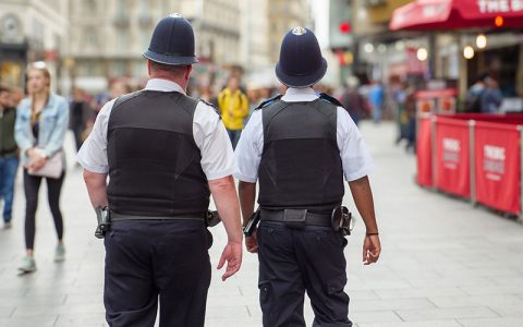 The image shows two police officers walking away, wearing uniforms and hats, amidst a bustling city street filled with people. Their presence suggests a focus on public safety.