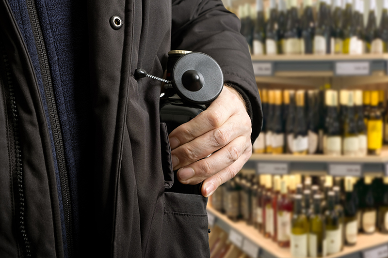 Close up of a mans hand and coat pocket as he attempts to steal a bottle of alcohol equipped with a Magnetic RF Bottle Tag security device, suggesting shoplifting