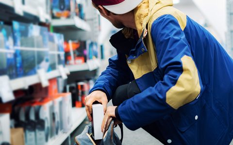 A person in a brightly colored jacket is crouching in a store aisle, discreetly placing an item into a backpack, suggesting a potential shoplifting incident.
