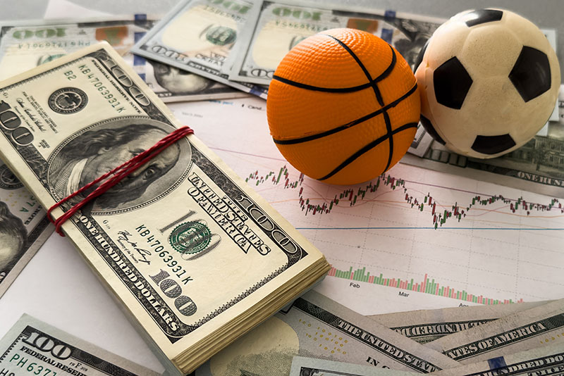 A football and basketball sitting on top of betting charts and cash, signifying online gambling and match fixing