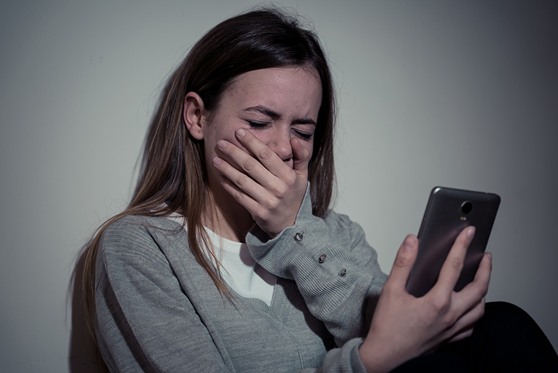 Sad desperate young teenager female girl on smart phone suffering from online bullying, sextortion, harassment felling lonely and hopeless sitting on bed at night.