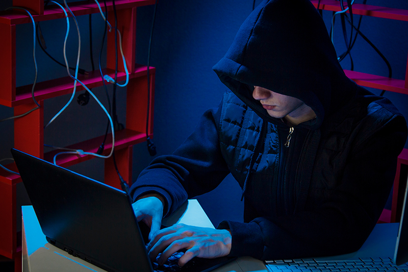 A teenager in a hoodie, face partially obscured, sits in a dark room with a black laptop
