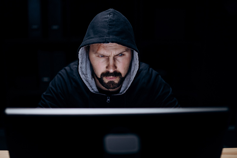 Bearded caucasian man in black hoodie working in a dark room on modern pc – possible terrorist or criminal