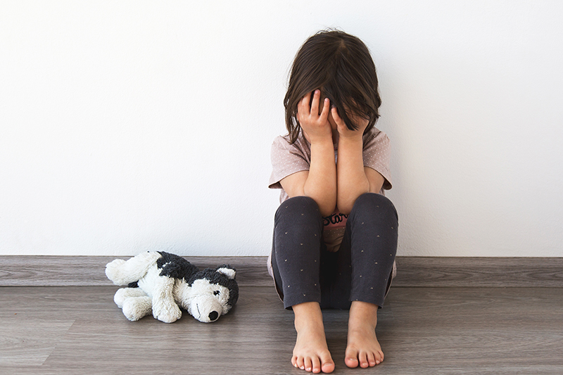 Little child sits on the floor against white wall, covering her face with her hands, child victim