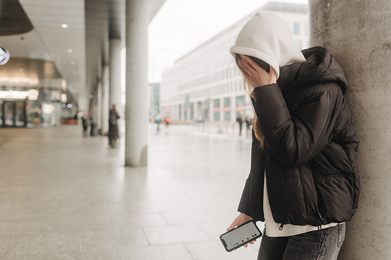 A young girl, wearing a hooded jacket, stands against a column in a public space, covering their face with one hand while holding a smartphone in the other.