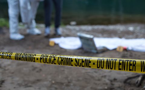 The image shows a police crime scene with yellow tape labeled "WARNING - POLICE CRIME SCENE - DO NOT ENTER," surrounding investigators examining a body wrapped in a white sheet.