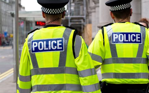The image shows two police officers in bright yellow vests with "POLICE" printed on the back, facing away from the viewer. They represent authority and public safety.