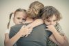 A mother (viewed from behind) hugging her two young children who look worried