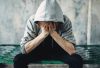 Man sitting on a bed frame, head in hands, wearing hoodie, face obscured