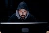 Bearded caucasian man in black hoodie working in a dark room on modern pc – possible terrorist or criminal