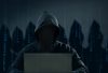 Hooded hacker sitting in a dark room at a laptop, with the silhouettes of other hackers behind him